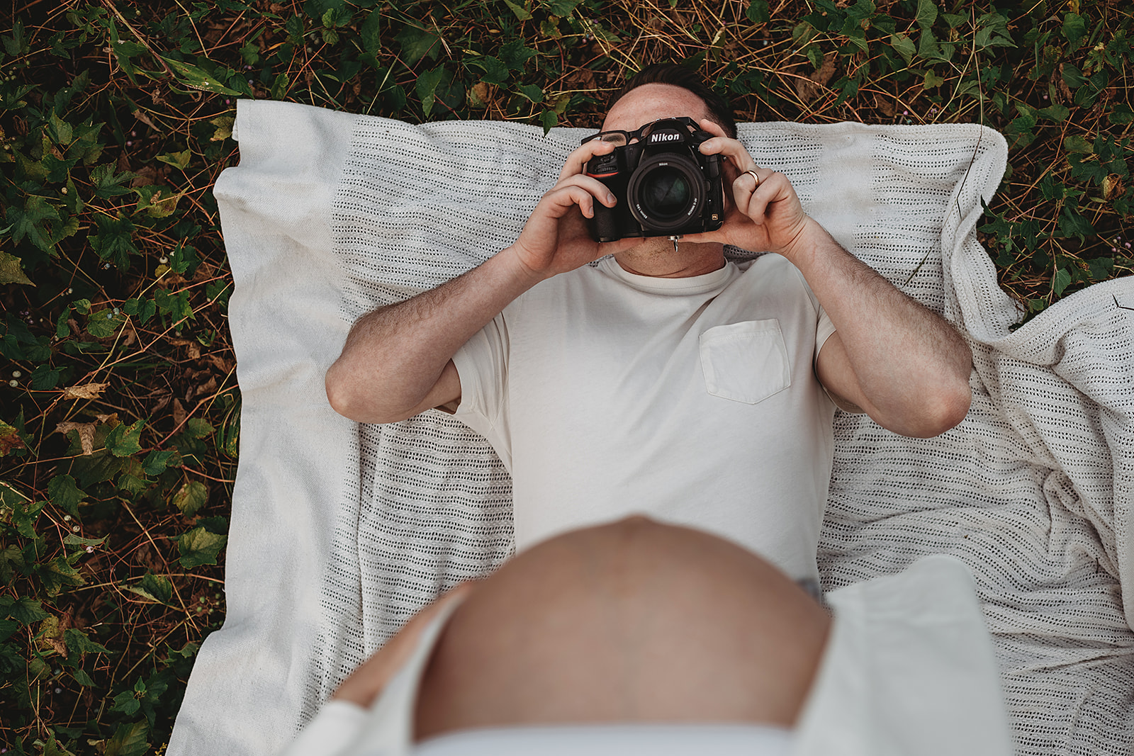 Man lying on a blanket in the grass during a maternity photoshoot in Baltimore, holding a Nikon camera and taking a photo of a pregnant woman’s belly in the foreground.